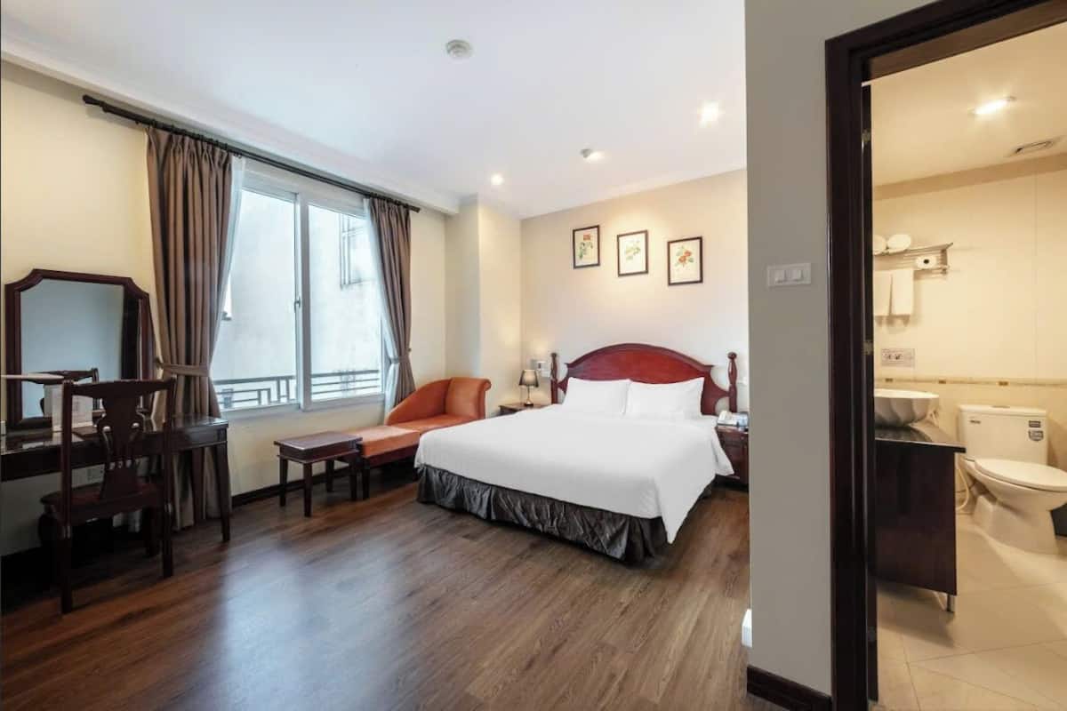 Habitació d'hotel amb un llit gran, una taula de treball amb una cadira, una taula petita i una finestra amb cortines.