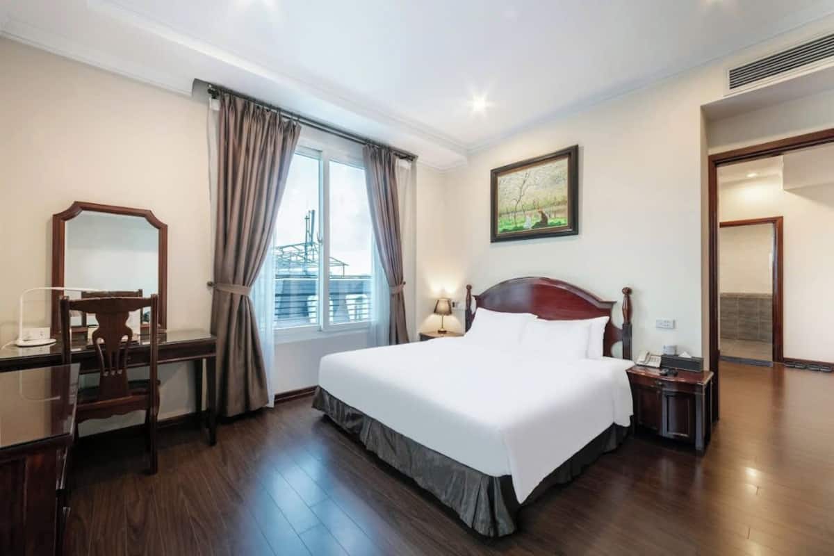Habitació d'hotel amb un llit gran, un escriptori amb cadira, una finestra amb cortines, una taula de nit amb làmpada i un quadre a la paret.