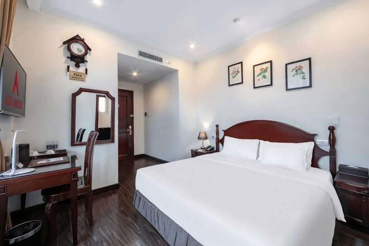 Habitació d'hotel amb un llit gran, una taula amb cadira, una làmpada de taula, un rellotge i quadres a la paret.