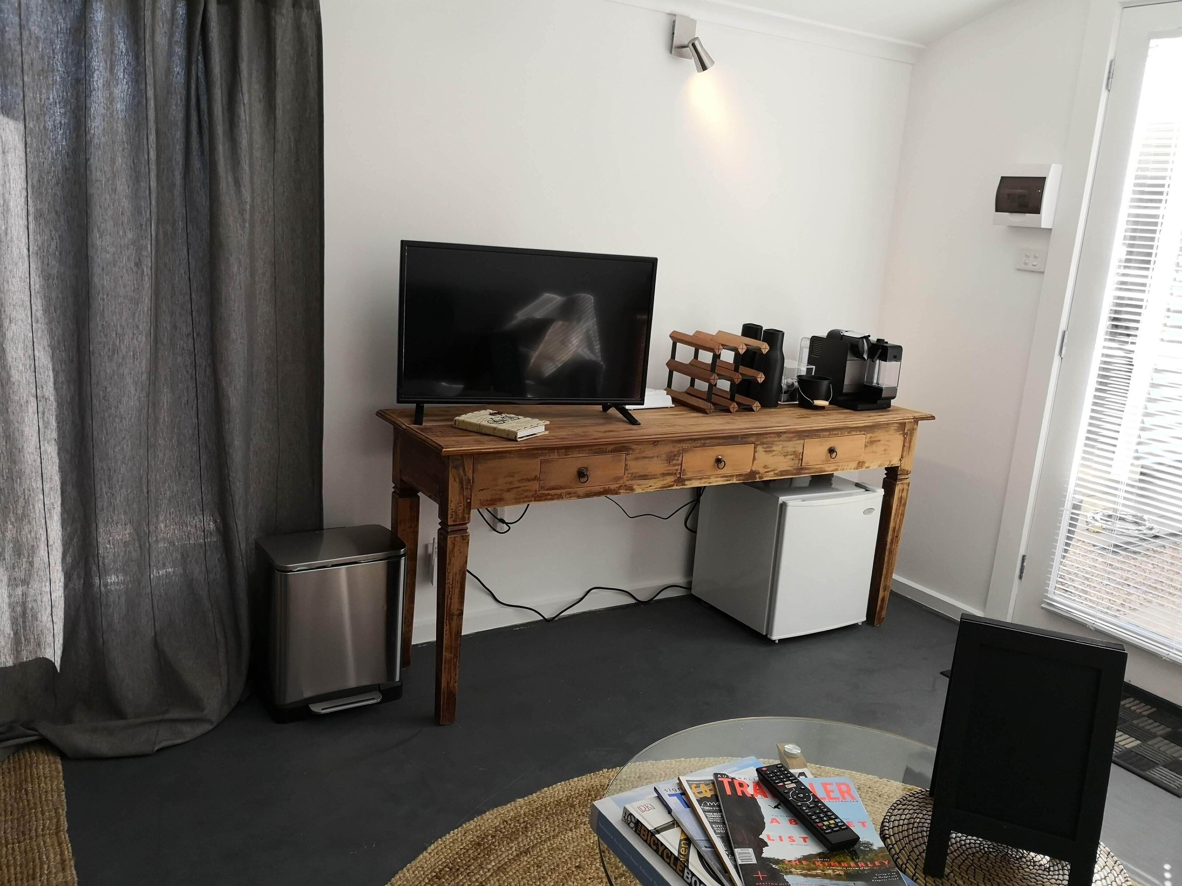 Een woonkamer met een houten bureau, een televisie, een kleine koelkast en een glazen salontafel met tijdschriften en afstandsbedieningen.