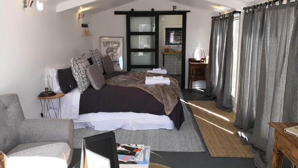 Un dormitorio con una cama grande, una mesita de noche con lámpara, una silla y vista al baño a través de una puerta de cristal.