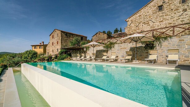 Una piscina con sillas de descanso y sombrillas, rodeada de edificios de piedra.