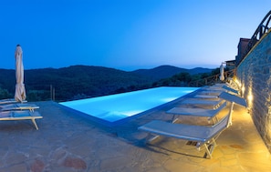 A rooftop infinity pool with lounge chairs and umbrellas, surrounded by stone walls and a view of the hills.
