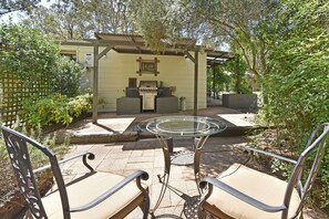 En uteplats med ett bord med glasskiva, två bekväma stolar, en grill och en pergola med en inramad tavla.