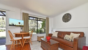 A living room with a wooden table, chairs, a brown leather sofa, a red colander, and a wall clock. - Olive Grove 1 - Your Perfect Hunter Valley Wedding Stay & Wine Country Escape (Pokolbin)