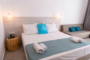 A hotel room with a bed, a bedside table with a phone, a sofa, and a blue pillow with text. - Lambi Resort (Kos)
