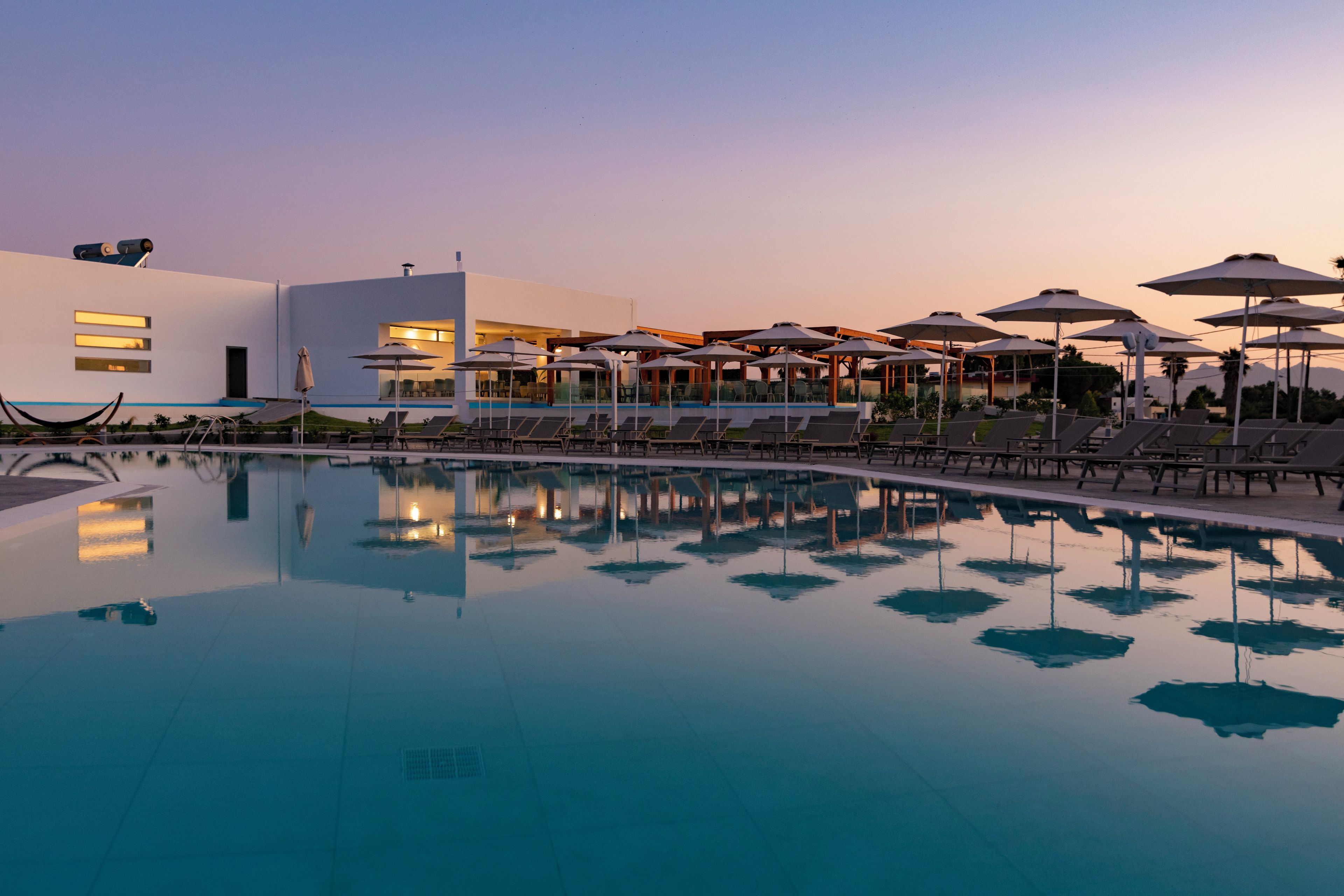 Area piscina con lettini e ombrelloni, un edificio moderno con ampie finestre e cielo sereno.