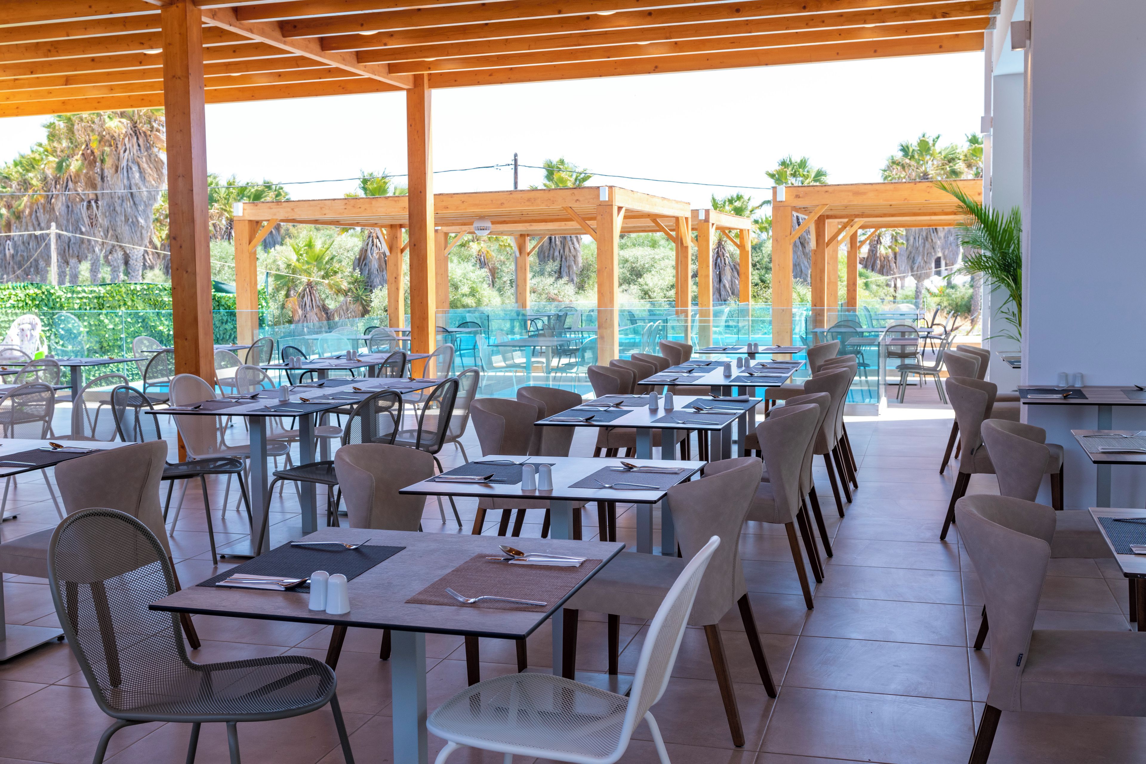 An outdoor dining area with multiple tables and chairs, a pool in the background, and a wooden pergola structure.