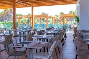 Un área de comedor al aire libre con varias mesas y sillas, una piscina al fondo y una estructura de pérgola de madera.