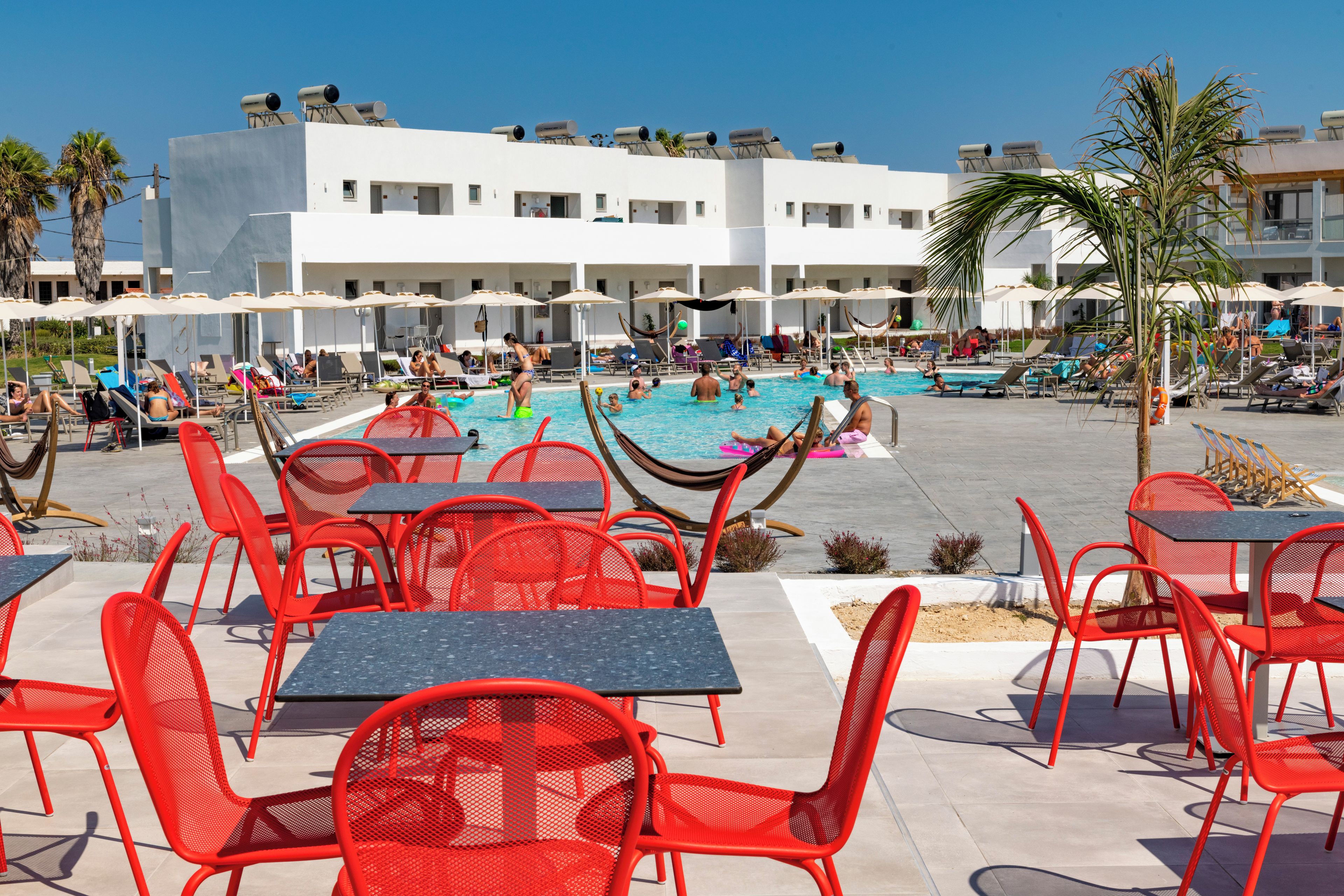 Un espace aménagé au bord de la piscine, avec des chaises et des tables rouges, une piscine et un bâtiment blanc doté de nombreux balcons.