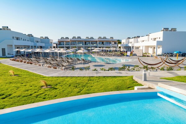 A pool area with lounge chairs and umbrellas, surrounded by modern buildings with balconies. - Lambi Resort (Kos)