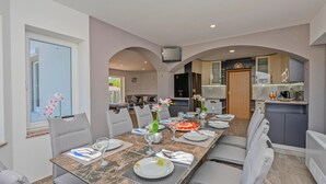 Un comedor con una mesa larga dispuesta para seis personas, flanqueada por dos sofás. Se ve una cocina al fondo con electrodomésticos modernos.