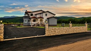 Una casa de dos pisos con un muro de piedra y un portón negro, rodeada por una cerca y un camino de acceso.