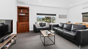 A modern living room with a large flat-screen TV, a wooden bookshelf, two leather sofas, a coffee table, and a window with a view of greenery.