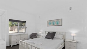A white bedroom with a bed, bedside tables, a chair, and a window.
