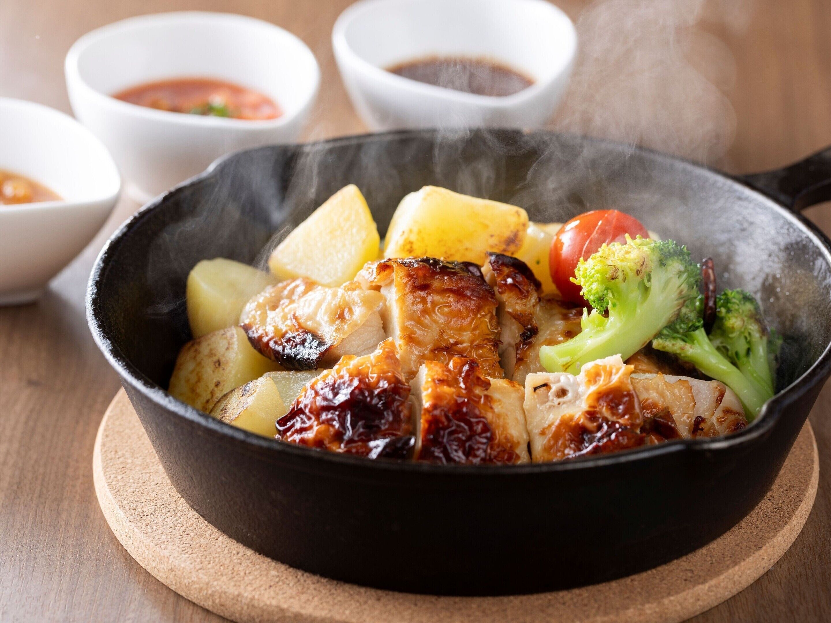 A skillet with grilled chicken, potatoes, and broccoli on a wooden board.