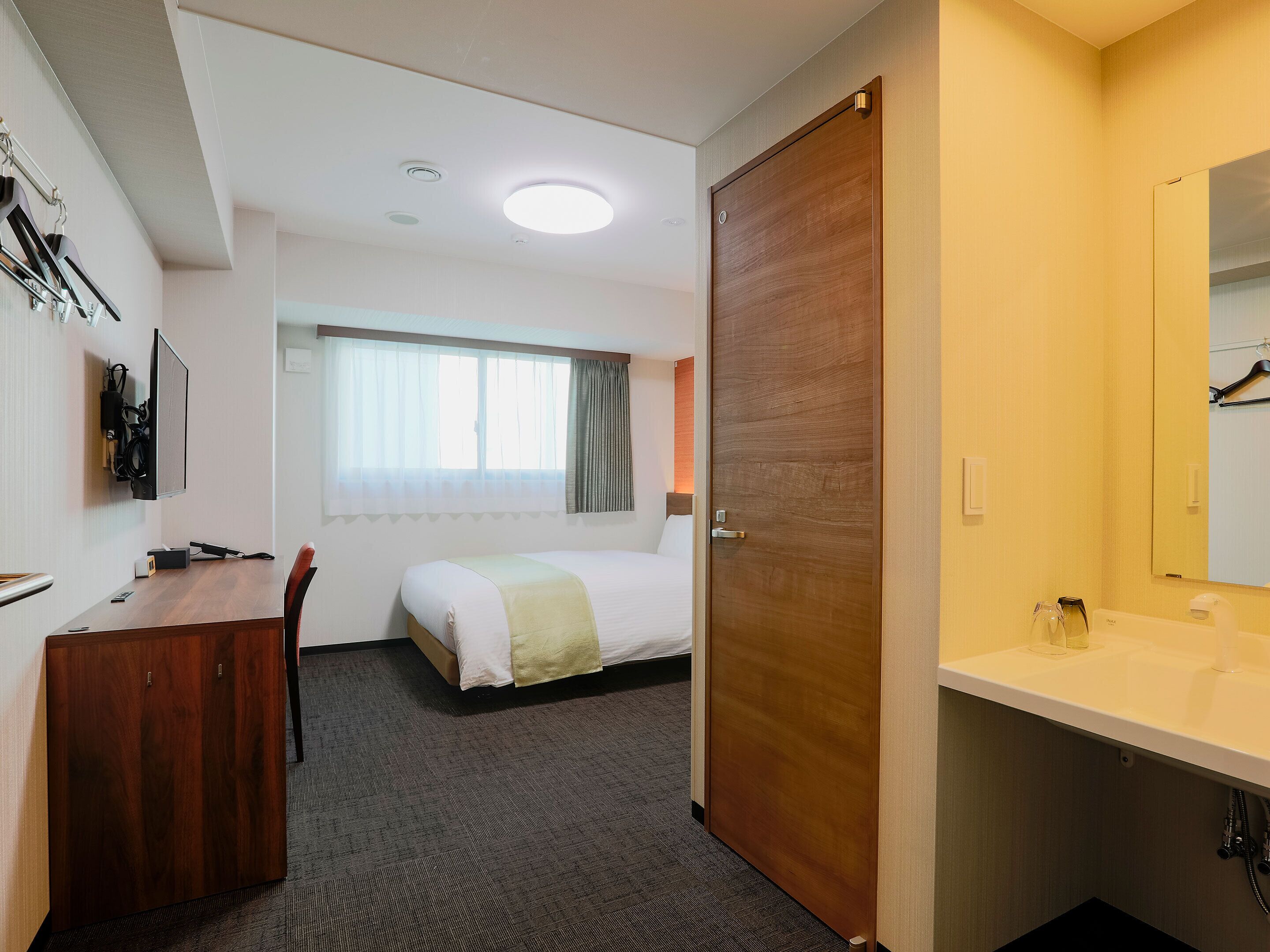 A hotel room with a bed, a desk, a TV, a window with curtains, a wooden door, and a bathroom with a sink and mirror.
