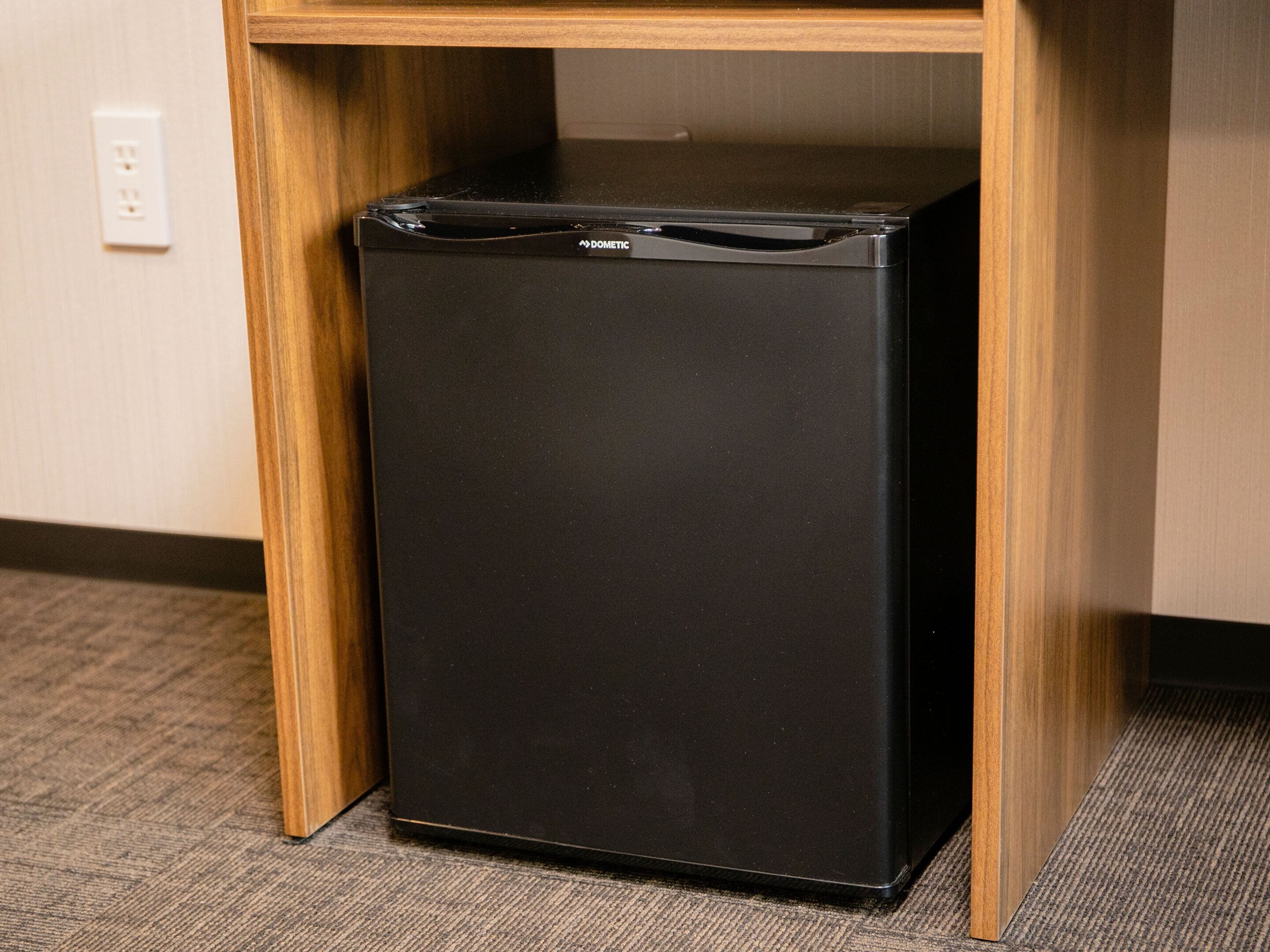 Un refrigerador negro debajo de un escritorio de madera en una habitación con piso alfombrado y un enchufe de pared.