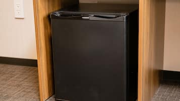 A black refrigerator under a wooden desk in a room with a carpeted floor and a wall socket.