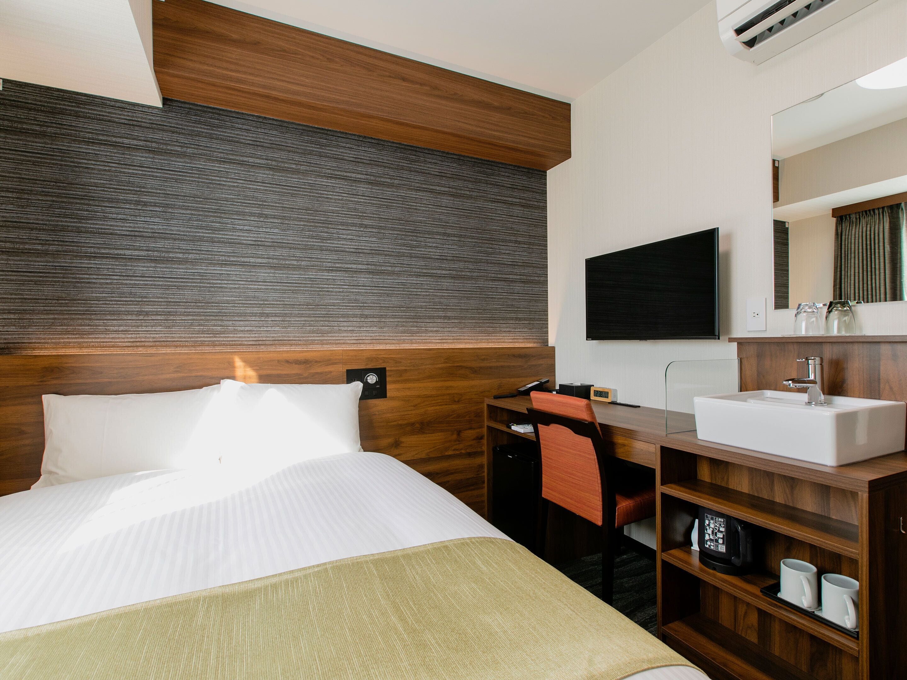 A hotel room with a bed, a desk, a chair, a TV, a sink, and a mirror.