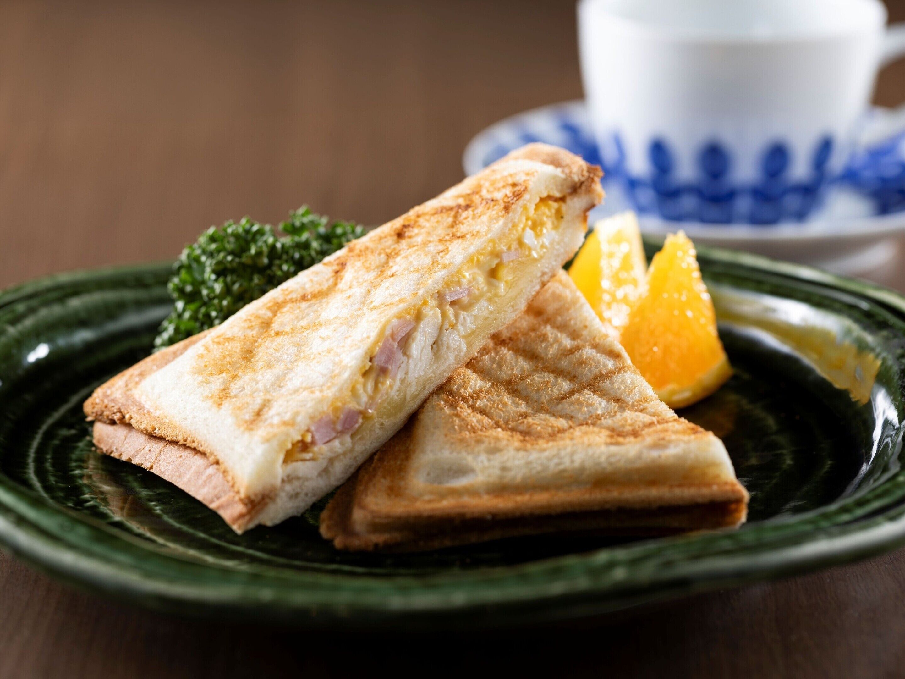A sandwich on a plate with a cup and saucer in the background.