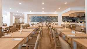 A modern dining area with long tables and chairs, a kitchen in the background, and a stone wall feature. - Hotel Mariant Park (Sant Llorenc des Cardassar)