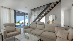 Ein modernes Wohnzimmer mit einem großen, beigefarbenen Ecksofa, einem Glastisch und einer Treppe, die zu einem Außenbereich mit Pool führt.