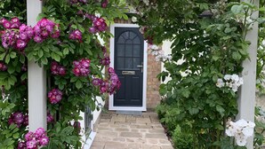 En stenlagd gång leder fram till en svart dörr, som omges av en träpergola med rosa blommor.