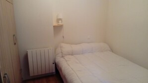 A small, single-bed room with a white bedspread, a bedside table with a lamp, and a radiator.