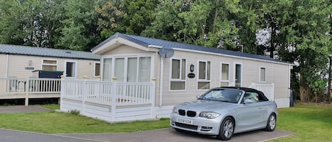 Un'abitazione mobile con rivestimento bianco e un balcone, parcheggiata di fronte a una zona boschiva.