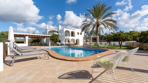 Uma piscina com espreguiçadeiras, um pergolado e um edifício branco com arcos ao fundo.