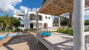 Uma área à beira da piscina com um pérgola, uma espreguiçadeira com uma toalha azul e uma piscina.