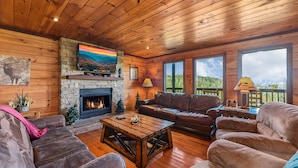 A cozy cabin interior with a stone fireplace, leather sofas, a wooden coffee table, and a television mounted on the wall. - Summer Break! Hot Tub | Views | 4-Story MTN Cabin (Gatlinburg)