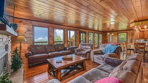 A cozy cabin interior with wooden floors, leather sofas, a fireplace, and large windows offering views of the outdoors. - Summer Break! Hot Tub | Views | 4-Story MTN Cabin (Gatlinburg)