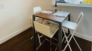 Een eetgedeelte met een tafel gedekt voor twee personen, een raam met uitzicht op huizen en een plafondlamp.