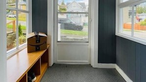 Een gang met een donkere muur, een witte deur en een raam. Er staat een houten bankje met schoenen en een pakket erop.