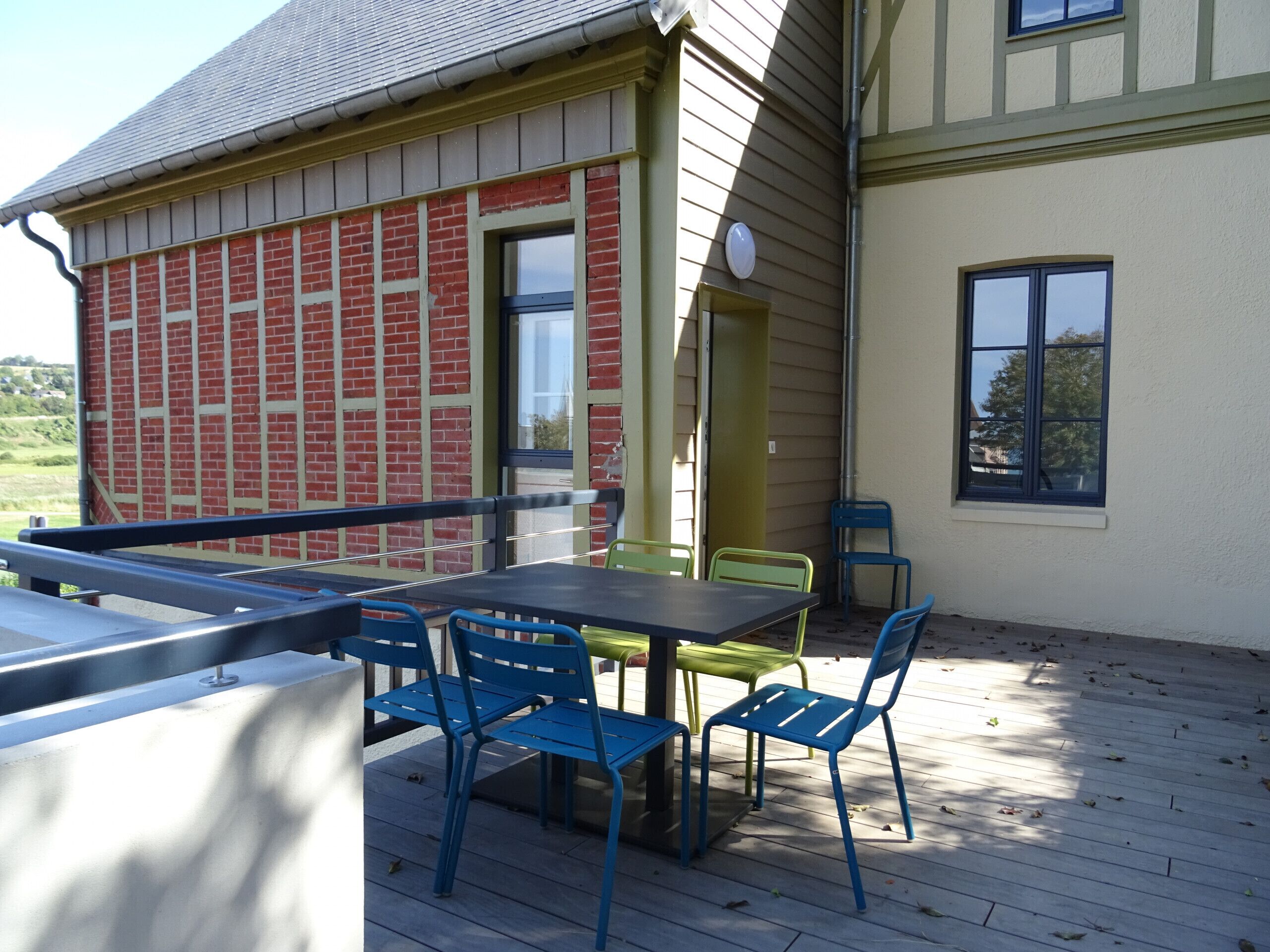 A covered outdoor patio with a table and chairs, a railing, and a building with a brick facade and a window.