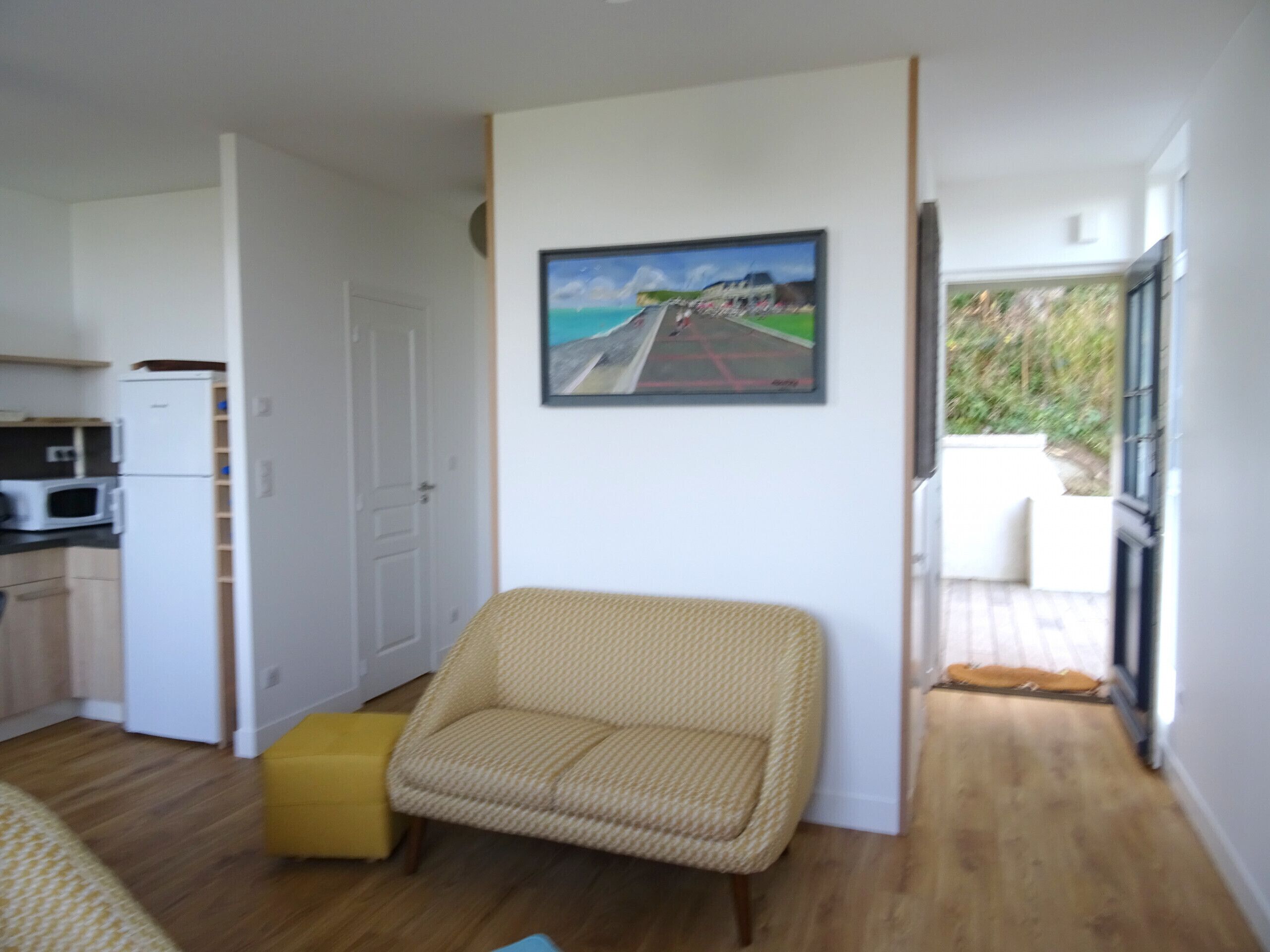 Een compacte, open woonruimte met een keukenhoek, een geel poefje, een rieten stoel, en een schilderij van een strandgezicht.