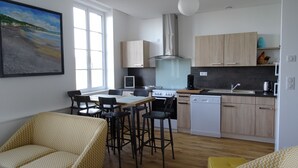 Une cuisine moderne avec des meubles en bois, une cuisinière à gaz et un lave-vaisselle. On y trouve deux chaises de salle à manger et une table. Un tableau représentant une plage est accroché au mur.