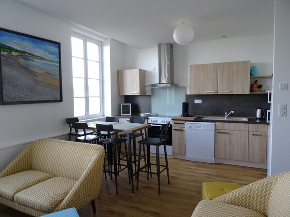 A modern kitchen with wooden cabinets, a gas stove, and a dishwasher. There are two dining chairs and a table. A painting of a beach is on the wall. - L'Horizon, duplex with terrace, sea vue (Hautot-sur-Mer)