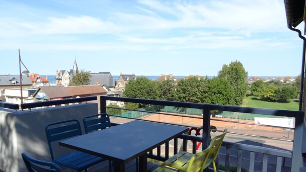 Un balcon avec une table et des chaises, donnant sur un court de tennis et des immeubles d’habitation.