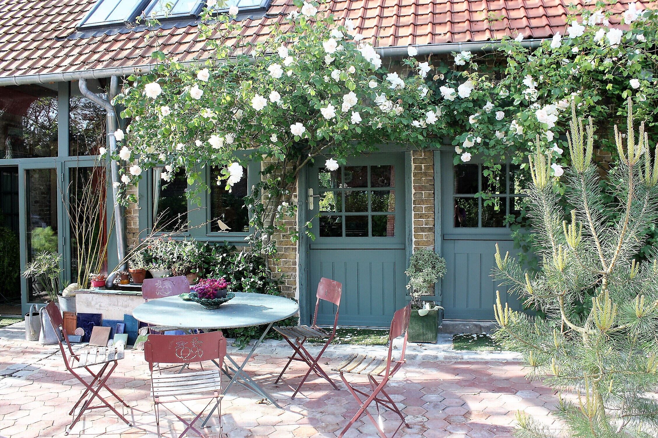 A patio with a round table and folding chairs, a brick pathway, and a house with a green door and windows.