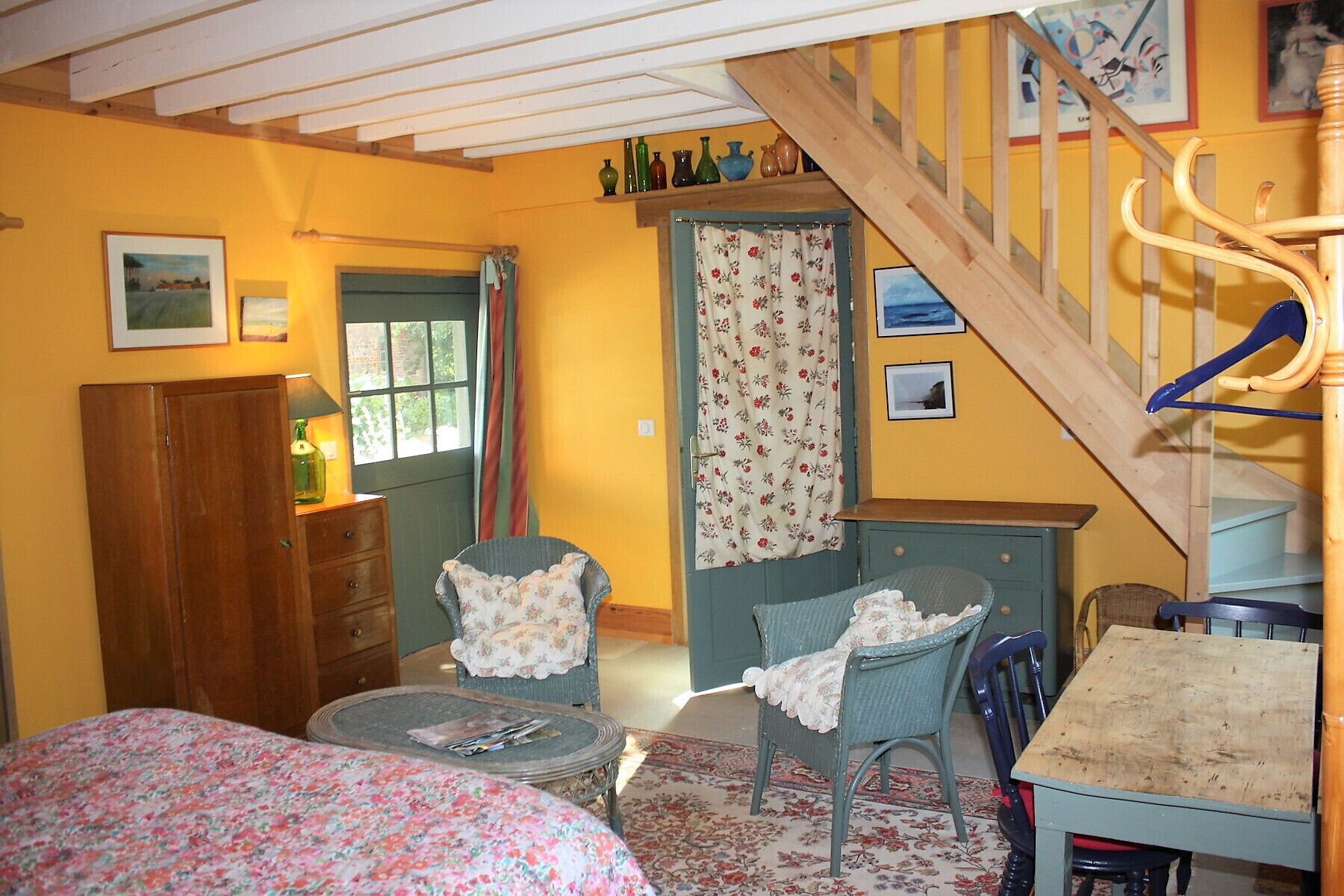 A cozy room with a wooden staircase, a bed with floral bedding, a chair with a floral cushion, a small table with magazines, and a wooden cabinet.