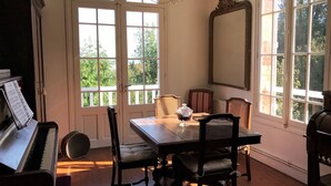 A dining area with a piano, a chandelier, and large windows with a view of greenery. - Roses,turn of the century house 100 meters from the sea (Hautot-sur-Mer)