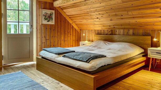 Un dormitorio de madera con una cama, una mesita de noche, una lámpara y un ventanal.
