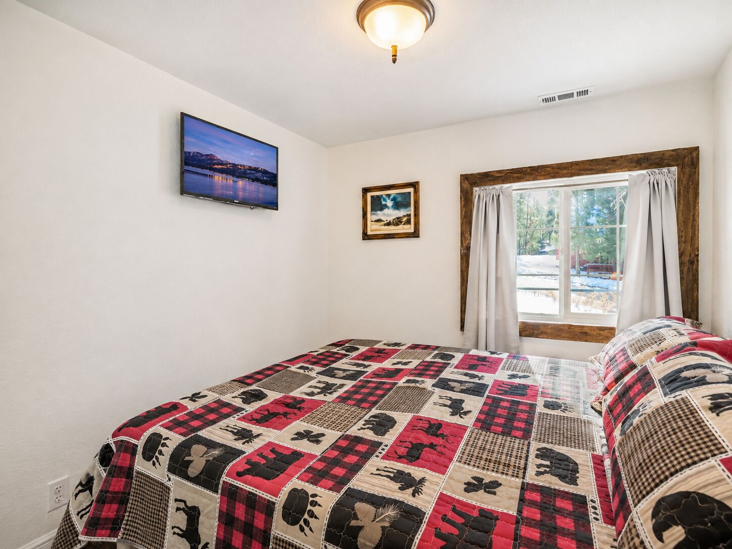 Un dormitorio con una cama, una ventana con cortinas, un televisor montado en la pared y un cuadro enmarcado.