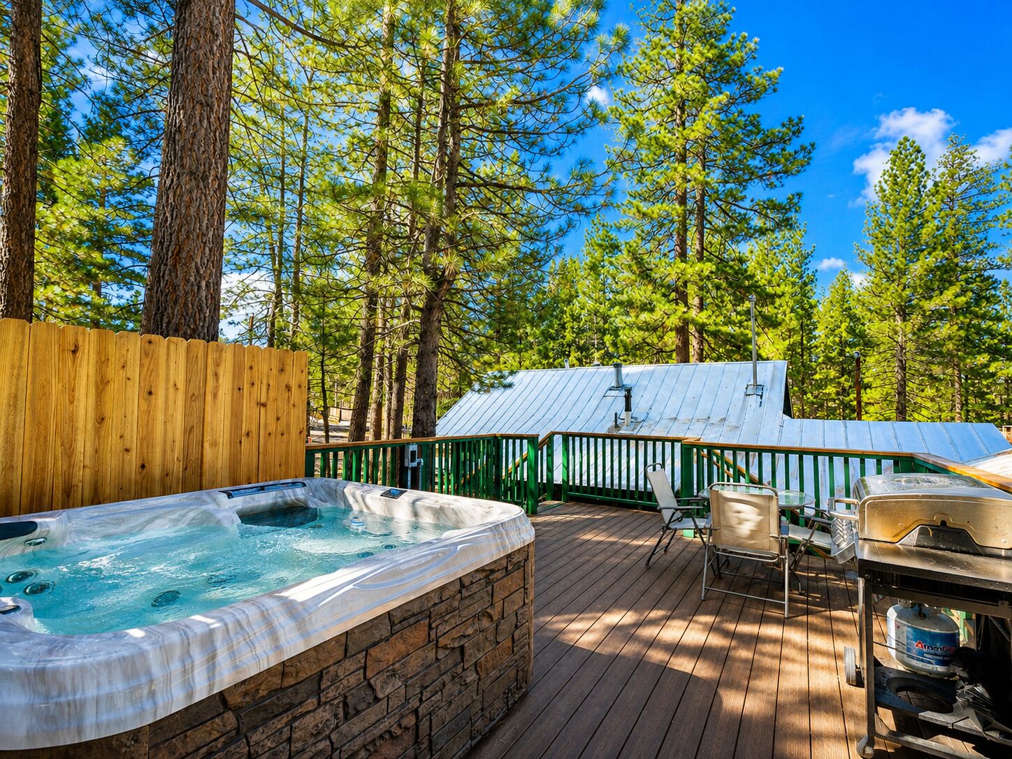 A hot tub on a wooden deck surrounded by trees, with a barbecue grill and a covered area in the background.
