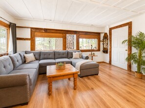 A cozy living room with a sectional sofa, a wooden coffee table, and a large window with a view of trees.