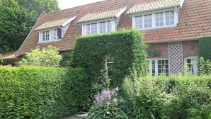 Uma casa de dois andares com telhado de telhas vermelhas, janelas emolduradas de branco e um jardim bem cuidado, com diversas plantas e um caminho de cascalho.
