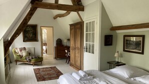 Um quarto com teto alto, vigas de madeira expostas, uma cama grande com roupa de cama branca, uma poltrona verde, um guarda-roupa de madeira e um quadro na parede.